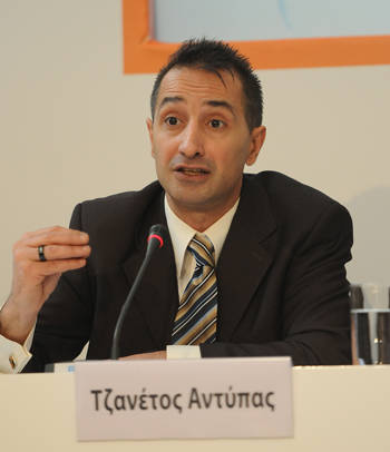 A man speaks into the microphone waving his hand and in front of him is a sign that reads Tzanetos Antipas